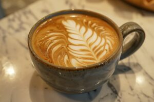 An up-close photo focusing on a cappuccino in a sleek, ceramic cup, showcasing the barista's skillful latte art - a delicate fern leaf design atop the frothy milk, set on the café's marble countertop